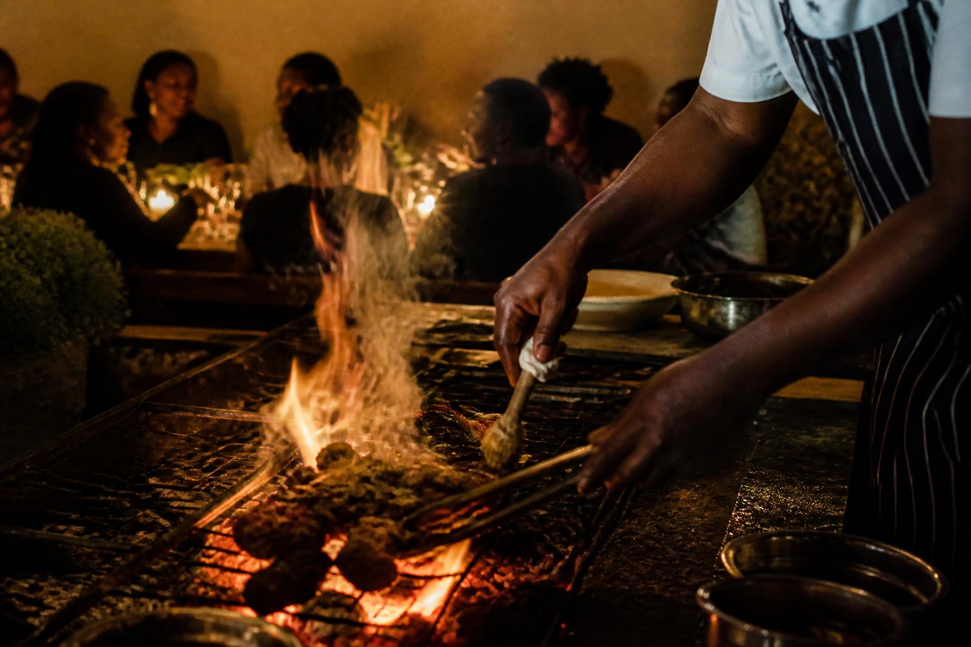 A cook works over an open flame while guests talk at a candlelit table behind the fire