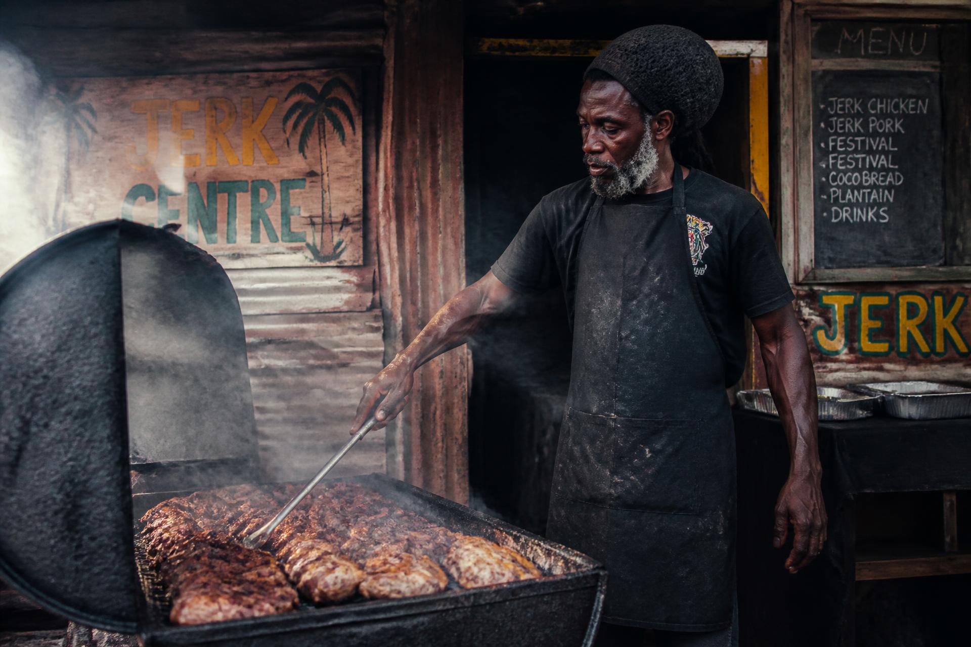 A cook turns meat over a smoking oil-drum jerk pit beside hand-painted signage
